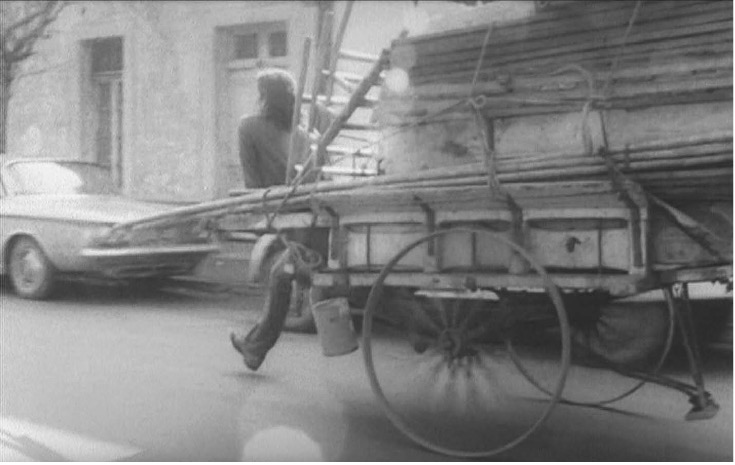 A black-and-white image depicting a person riding on the back of a cart carrying wood.